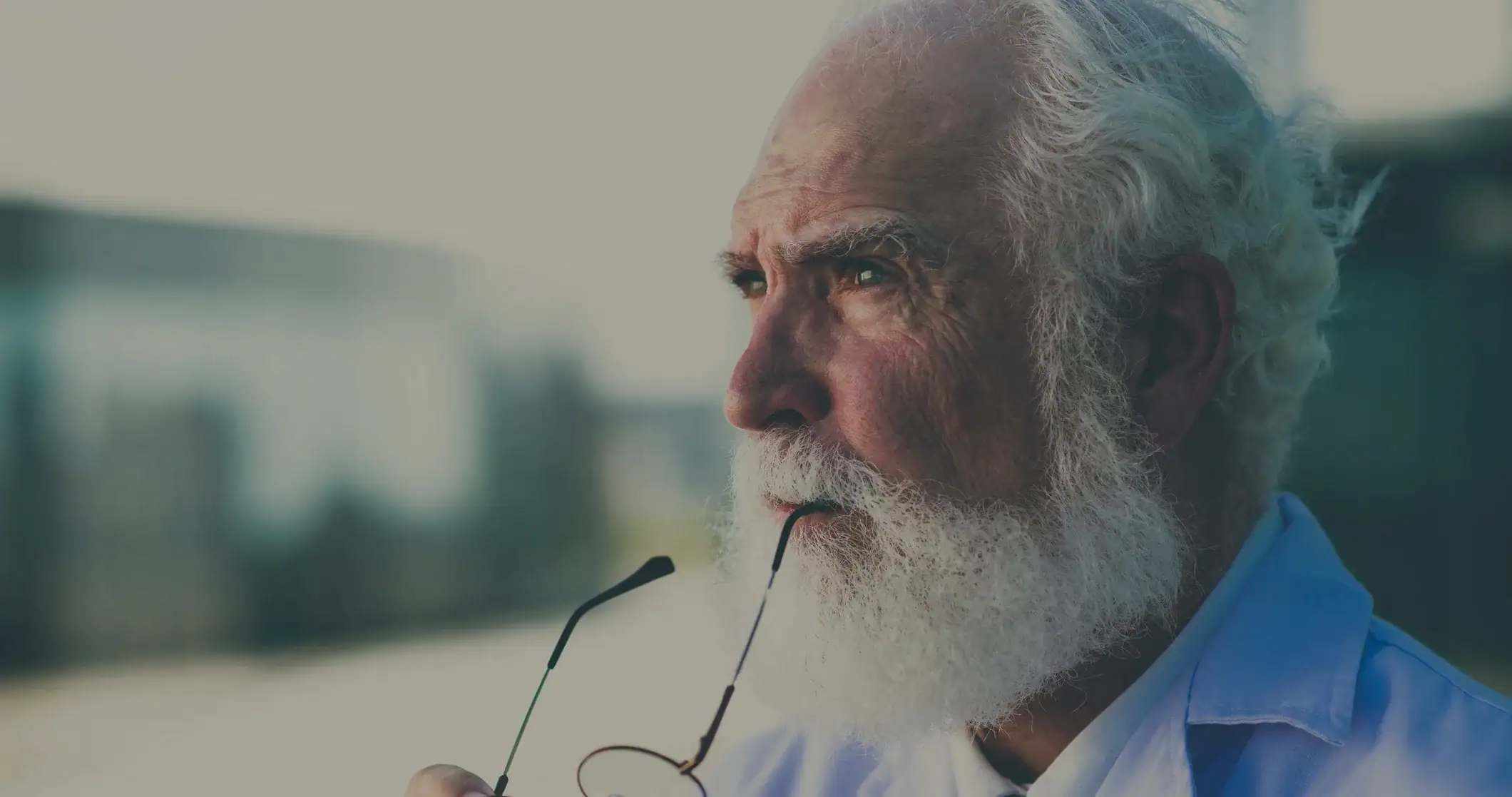 An elderly man with white hair and beard, holding glasses, looking thoughtfully into the distance.
