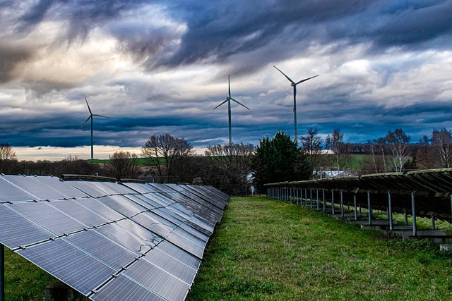 Solar panels and wind turbines in a renewable energy farm under cloudy sky.