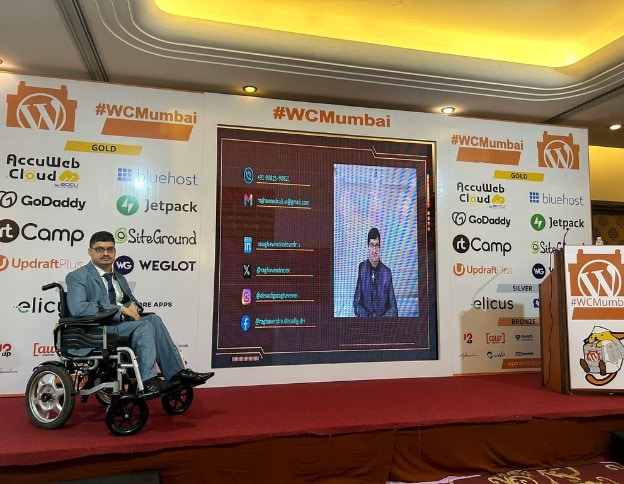 An individual in a wheelchair presenting at a tech conference with a large screen and sponsor banner.