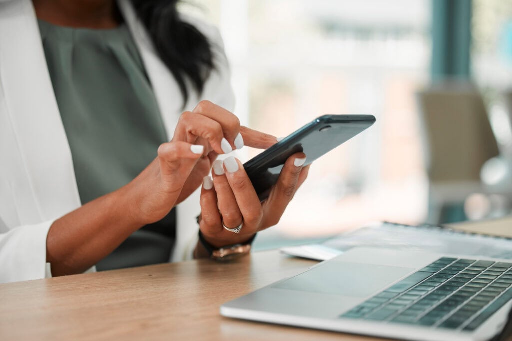 Woman using smartphone with laptop on desk, tech workspace.
